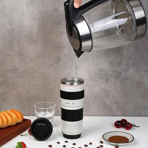A clear electric kettle pours water into a white travel mug designed to resemble a Canon camera lens. The mug has black accents and a stainless steel interior. On the table are coffee beans, coffee grounds in a dish, a glass of water, a pastry on a wooden board, and the mug's black lid labeled "Caniam."kitchen supply store, Kitchen items, coffee mug,travel essentials, travel accessories