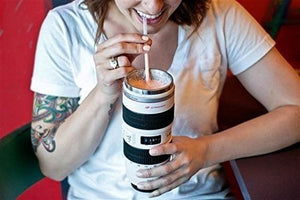A smiling woman wearing a white t-shirt and a tattoo on her left arm is drinking from a white travel mug designed to look like a camera lens. She is using a straw, and the mug has black accents and a stainless steel rim. kitchen supply store, Kitchen items, coffee mug,travel essentials, travel accessories