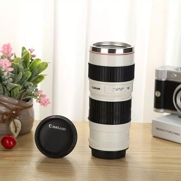 A Canon lens-shaped travel mug, resembling a camera lens, stands on a wooden surface with its black lid beside it. The mug has white and black sections with printed details. A potted plant and a miniature camera are visible in the background.kitchen supply store, Kitchen items, coffee mug, travel essentials, travel accessories