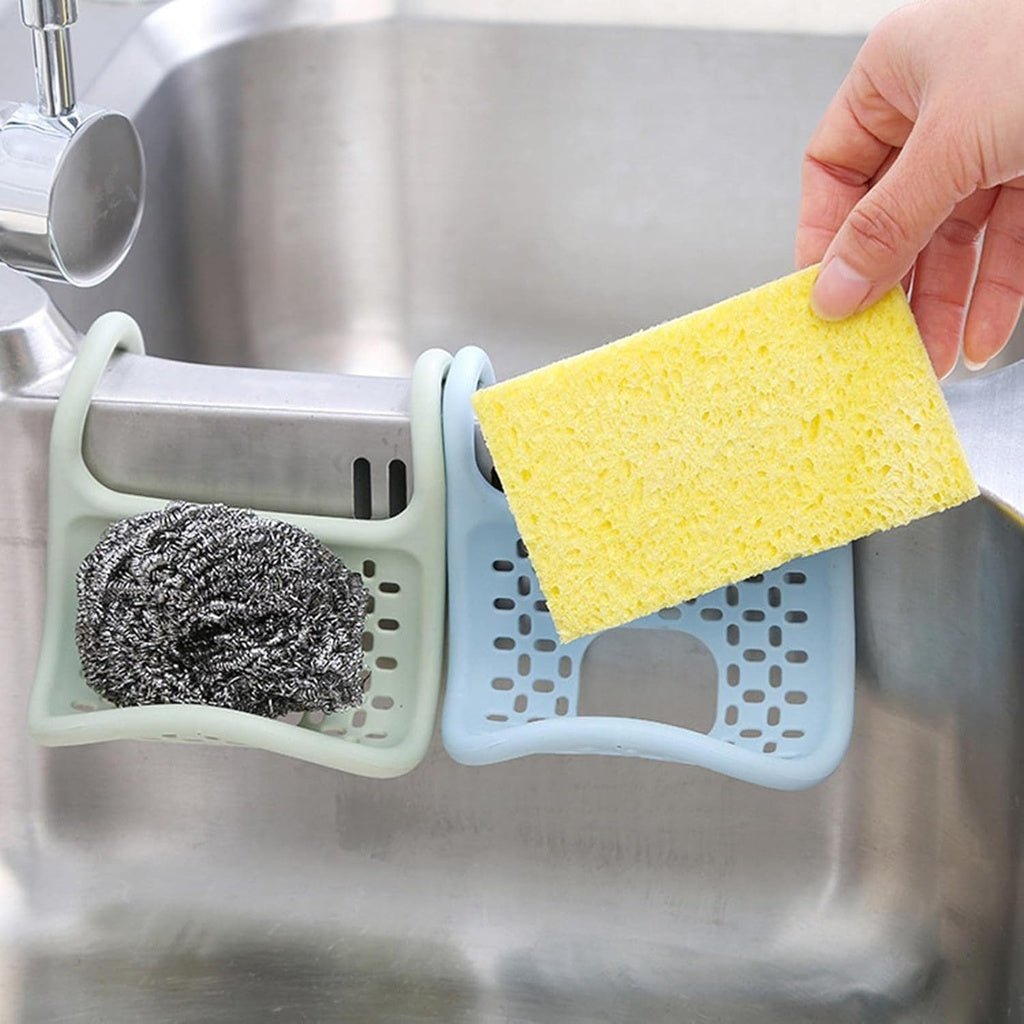 A hand places a yellow kitchen sponge into a light blue, foldable silicone sink caddy that is hanging over the edge of a stainless steel kitchen sink. Another similar caddy in light green hangs beside it, holding a metal scouring pad.sink caddy, sponge holder, sink organiser, kitchen organiser, dish sponge holder, silicone caddy, foldable caddy, kitchen accessories, household items, kitchen items.