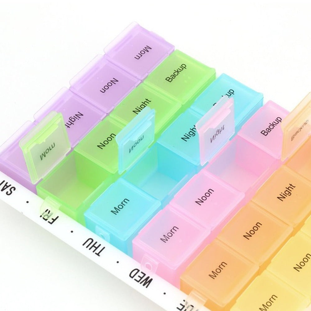 Close-up of a multi-compartment pill organizer with translucent, pastel-colored sections labeled for different times of the day ("Morn," "Noon," "Night," "Backup") and days of the week (partially visible "SAT," "FRI," "THU," "WED").