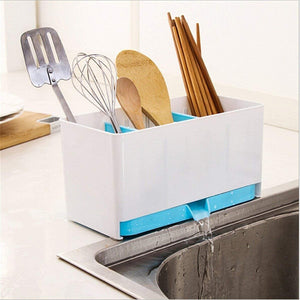 A white and blue plastic kitchen utensil holder with three compartments sits on the edge of a stainless steel sink. The holder contains various kitchen tools including a metal spatula, a wire whisk, wooden spoons, and chopsticks. Water is dripping from the bottom of the holder into the sink.utensil holder, kitchen organiser, cutlery drainer, spoon holder, chopsticks holder, kitchen storage, dish drying rack, kitchen accessories, household items, kitchen items.