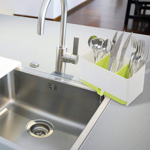 A white and light green plastic multipurpose kitchen rack sits on the edge of a stainless steel kitchen sink, next to a modern chrome faucet. The rack has three compartments and holds various silverware, including spoons, forks, and knives.utensil holder, kitchen organiser, cutlery drainer, spoon holder, fork holder, knife holder, sponge basket, kitchen storage, dish drying rack, kitchen accessories, household items, kitchen items,