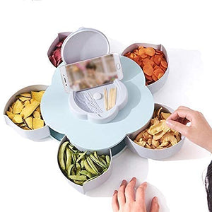 A light blue flower-shaped rotating tray with snacks in compartments and a phone holder in the center. Hands reach for snacks.snack tray, phone holder, rotating, serving dish.