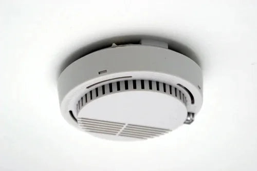 A white, round smoke detector is mounted on a white ceiling, viewed from below. The device has vertical and horizontal vents visible on its surface. smoke detector, ceiling mounted, fire safety, home safety, safety device, household items, home essentials.