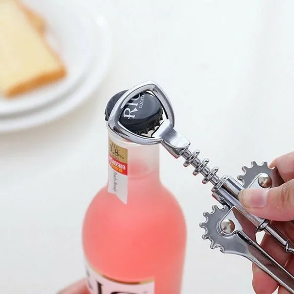 Two hands are using a silver wing corkscrew to open a bottle of red wine. The cork is partially extracted from the bottle. The background is blurred, suggesting a home setting. Wine opener, corkscrew, bottle opener, barware, kitchen tool, home essential.