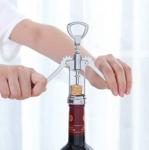 Two hands are using a silver wing corkscrew to open a bottle of red wine. The cork is partially extracted from the bottle. The background is blurred, suggesting a home setting. Wine opener, corkscrew, bottle opener, barware, kitchen tool, home essential.