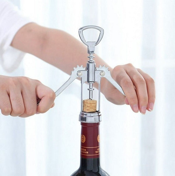 Two hands are using a silver wing corkscrew to open a bottle of red wine. The cork is partially extracted from the bottle. The background is blurred, suggesting a home setting. Wine opener, corkscrew, bottle opener, barware, kitchen tool, home essential.
