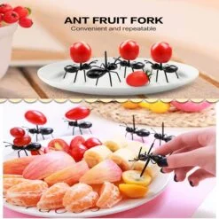 At the top, ant fruit fork is placed on an empty white plate. At the bottom, ant fruit fork is inserted in the slices of apple, oranges, etc. These mini fruit picks are of red and black color.