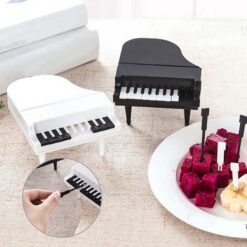 One piano fruit fork is black in color and another piano fruit fork is white in color. One plate filled with fruits are shown where piano fruit fork is being used.