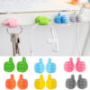 Different colors thumb hooks are shown. Pink self adhesive thumb hook is holding keys. Green self adhesive thumb hook is holding ear phones and white self adhesive thumb hook is holding a charging cable wire.