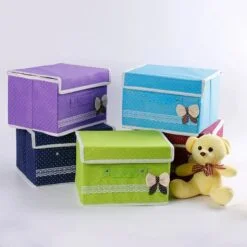 5 non woven storage box of different colors are kept on a table along with a teddy bear.