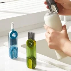 cup lid cleaning brush is presented in a blue and green color near wash basin. Also a lady is cleaning cream color bottle top with grey color cup lid cleaning brush.