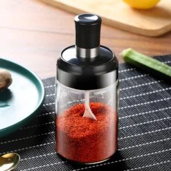 On a table a red chilli powder is filled in a glass condiment jar with lid and spoon.