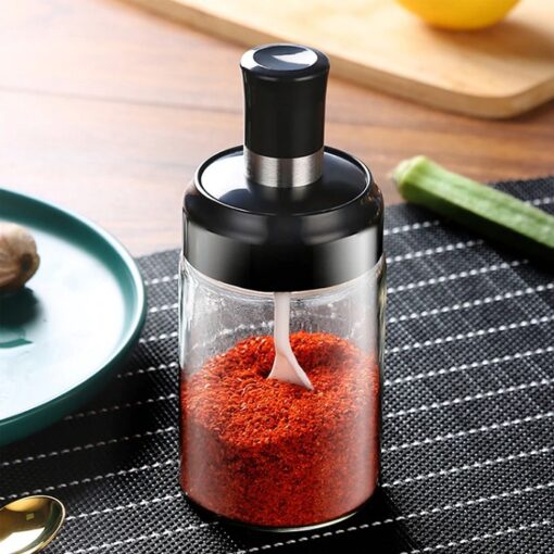 On a table a red chilli powder is filled in a glass condiment jar with lid and spoon.