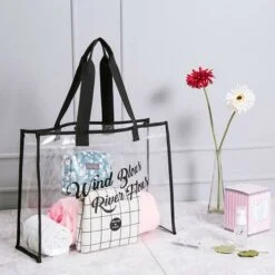 Transparent large clear tote bag is being placed on a white color table besides flowers.