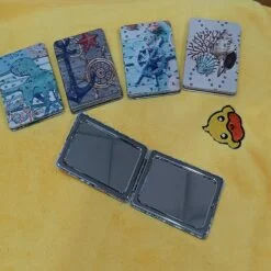 4 different designs and colors rectangular travel pocket mirror are closed and placed on a table. One mirror is wide open.