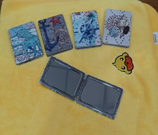 4 different designs and colors rectangular travel pocket mirror are closed and placed on a table. One mirror is wide open.