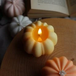 Orange and yellow color pumpkin shaped candles are kept on a table and white and pink color pumpkin shaped candles are kept under the table besides an open book.