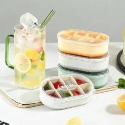 Ice cube tray with lid is shown in different colors besides a juice glass on the table