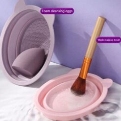 Sponge is being cleaned in a purple color makeup brush cleaning tray and makeup brush is being cleaned in a pink color makeup brush cleaning tray