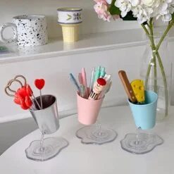 Bucket pen holder is presented in 3 different colors on a white color table besides flowers. This creative pencil holder is used to store knives, pens, pencils, etc.