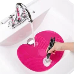 Woman is cleaning her makeup brush in a majenta color makeup cleaning mat placed in a washbasin