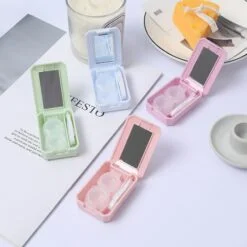 Contact lens case with mirror is shown in blue, green, purple, and pink color besides cheese candle, workbook, and a glass.
