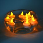 A set of 12 flameless LED Diwali Lights floating in a crystal clear bowl filled with water. The candles feature a realistic flame effect and are illuminated with a warm white glow, creating a calming and elegant atmosphere.