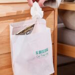 Close-up of a white self-adhesive trash bag dispenser mounted on a cabinet door, with a hand dispensing a trash bag. Multiple self-adhesive trash bag dispensers in various colors (depending on availability) neatly arranged on a shelf. A kitchen scene featuring a self-adhesive trash bag dispenser mounted under the sink, with a trash bag hanging out for easy access.